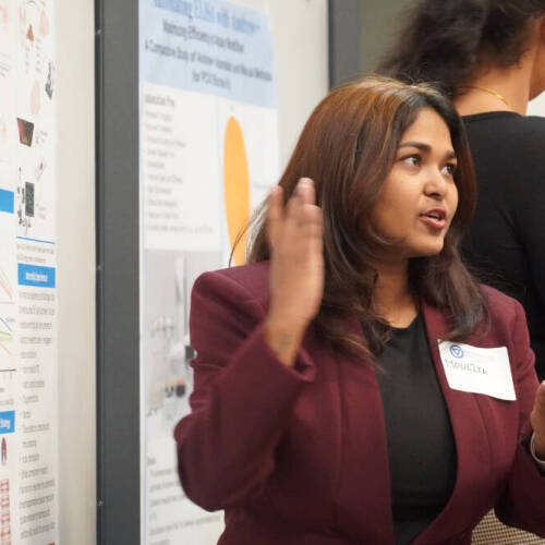 A woman wearing a maroon blazer and name tag labeled "Moulika" is presenting a scientific poster at a conference or academic event. She is speaking animatedly and using hand gestures to explain her work to others. with scientific posters in the background